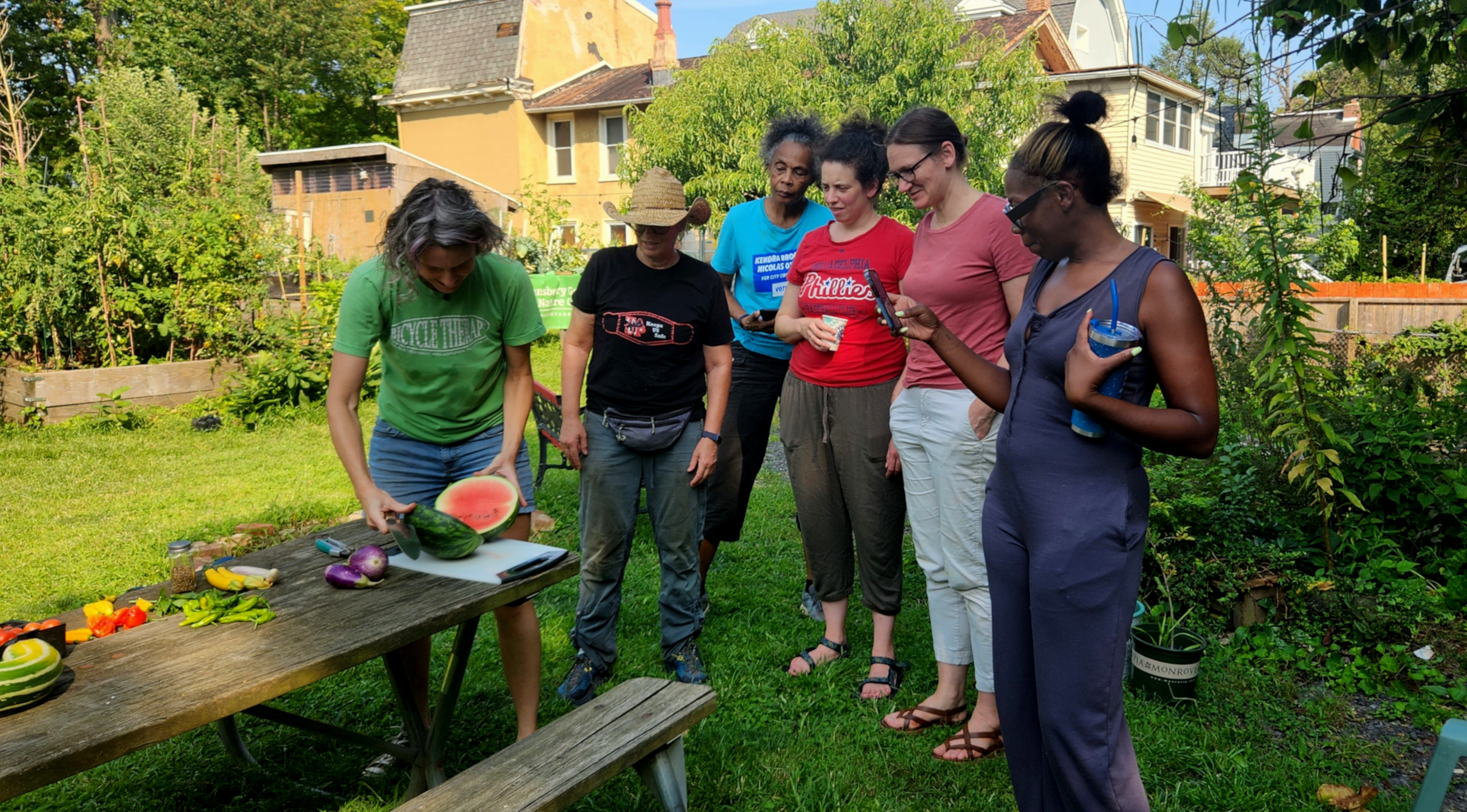 photo: group gathered around picnic table watching a woman cut open a watermelon